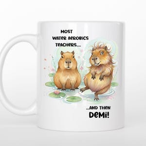 May include: White ceramic mug featuring two cartoon capybaras in a water aerobics scene. The mug has the text "MOST WATER AEROBICS TEACHERS....AND THEN DEMI!" in black. The capybaras are brown and tan, with a whimsical design.