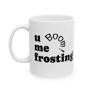 May include: White ceramic mug with the text "u me boom! frosting" in black, whimsical lettering. The mug has a curved handle and a cylindrical shape, suitable for hot or cold beverages.