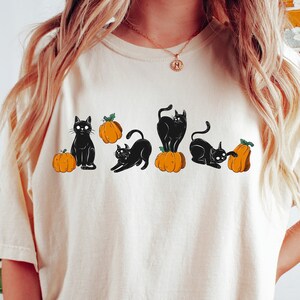 May include: A cream-coloured t-shirt with a graphic of black cats and orange pumpkins. The cats are in different poses, sitting, stretching, and playing. The t-shirt has a relaxed fit, suitable for casual wear.