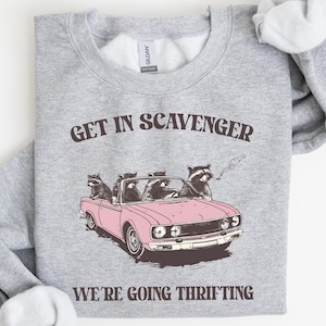 Op de afbeelding: Een grijs sweatshirt met een afbeelding van wasberen die een roze auto rijden. De tekst op het sweatshirt luidt "Get in Scavenger We're going thrifting."