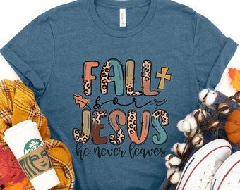 Könnte beinhalten: Ein blaues T-Shirt mit dem Text "Fall for Jesus he never leaves" in bunten Buchstaben mit Leopardenmuster. Das Shirt ist umgeben von herbstlichen Gegenständen wie einem karierten Schal, Kürbissen und Herbstblättern.