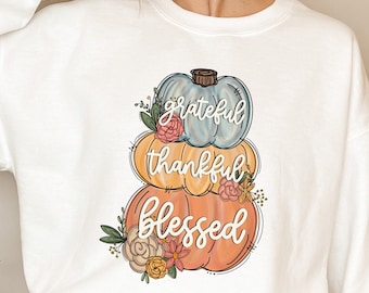 May include: White sweatshirt with a design of three stacked pumpkins in pastel shades of blue, yellow, and orange. The pumpkins have the words "grateful," "thankful," and "blessed" on them, with floral details.