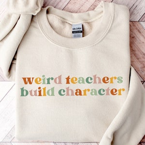 May include: A beige sweatshirt with the text "weird teachers build character" in colourful letters. The letters are in a retro style font.