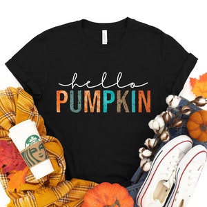 Peut inclure: T-shirt noir avec l'inscription "hello PUMPKIN" en blanc et en lettres colorées. Le t-shirt est entouré d'articles sur le thème de l'automne, dont une écharpe jaune, des citrouilles, des feuilles et des baskets blanches.