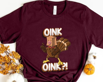 May include: A burgundy t-shirt featuring a cartoon turkey wearing a pig mask, with the words "OINK OINK?!" printed in white. The tee is a short-sleeved crew neck, suitable for everyday wear.