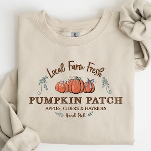 Może przedstawiać: Beżowa bluza z brązowym nadrukiem, na którym widnieje napis "Local Farm Fresh Pumpkin Patch Apples, Ciders & Hayrides Hand Picked". Nadruk zawiera również trzy dynie.