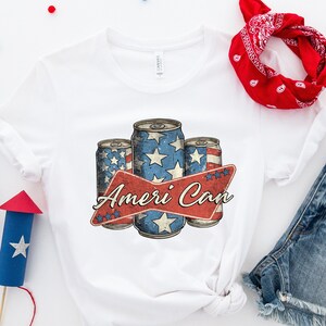 May include: White t-shirt with a graphic of three red, white, and blue cans with stars and the text "Ameri Can". The shirt is laid out on a white surface with a red bandana, blue denim shorts, and red and blue paper firecrackers.