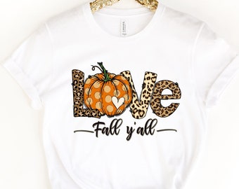 Puede incluir: Camiseta blanca con un diseño gráfico de la palabra "LOVE" con una calabaza en la "O". La calabaza es naranja con lunares blancos y un pequeño corazón blanco. Las letras están rodeadas de un estampado de leopardo. El texto "Fall y'all" está debajo del gráfico.