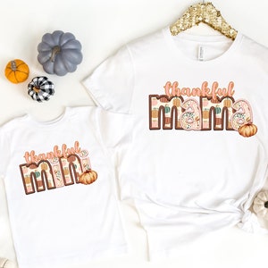 Pode incluir: Duas camisetas brancas com o texto "thankful mama" e "thankful mini" em letras marrons com um padrão de outono. As camisetas estão sobre um fundo branco com decorações de outono.