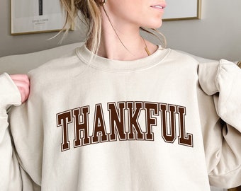 May include: Beige sweatshirt featuring the word "THANKFUL" in brown block lettering. The sweatshirt has a crew neck and long sleeves. The text is in a collegiate style font. The overall style is casual and comfortable.