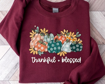 May include: A maroon sweatshirt with a seasonal design. The graphic shows a collection of pumpkins in different colours and patterns, with flowers, and the words "thankful + blessed" in white.