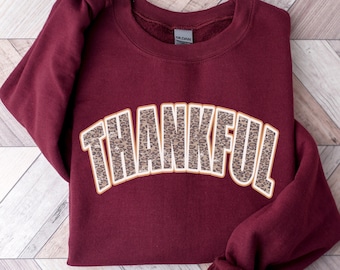 May include: A maroon sweatshirt featuring the word "THANKFUL" in a leopard print pattern. The text is arched and outlined in tan. The sweatshirt is folded on a light wood-grain surface.