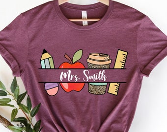 May include: Burgundy t-shirt with a graphic of a red apple, pencil, coffee cup, and ruler. The text "Mrs. Smith" is printed on the shirt.