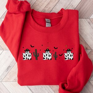 May include: A red sweatshirt featuring a Halloween-themed design. The design includes three ghosts wearing cowboy hats and cow-print outfits, a cactus, bats, and stars. The sweatshirt is made of a soft material.