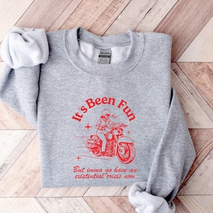 Puede incluir: Sudadera gris con un gráfico rojo de una persona conduciendo una motocicleta. El texto dice "It's Been Fun But imma go have an existential crisis now."