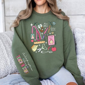 May include: Green sweatshirt with a colourful graphic design of school supplies, including a coffee cup, scissors, pencils, a ruler, a book, and an apple. The text "More coffee please" and "Lesson Plans" are included in the design. The text "Can't talk right now, doing teacher stuff" is on the sleeve.