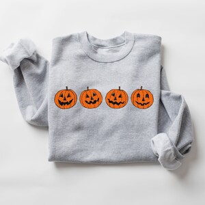 May include: A gray sweatshirt with a row of four orange jack-o'-lantern pumpkins with different facial expressions.
