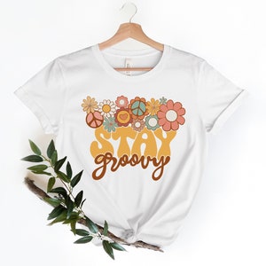 May include: White t-shirt with a yellow "Stay Groovy" graphic with brown lettering. The graphic is surrounded by colorful flowers and peace signs.