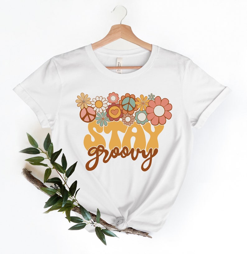 May include: White t-shirt with a yellow "Stay Groovy" graphic with brown lettering. The graphic is surrounded by colorful flowers and peace signs.