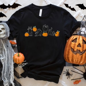 May include: Black t-shirt with a Halloween design showing black cats on orange pumpkins. The image includes Halloween decorations such as a jack-o'-lantern, spiders, and bat cutouts.