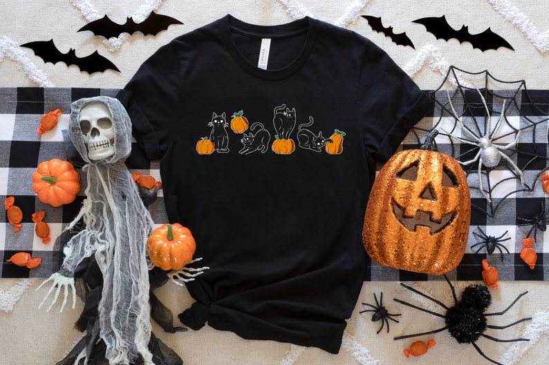 May include: Black t-shirt with a Halloween design showing black cats on orange pumpkins. The image includes Halloween decorations such as a jack-o'-lantern, spiders, and bat cutouts.
