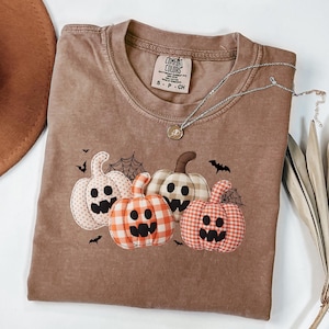 Peut inclure: Un t-shirt Comfort Colors marron avec quatre citrouilles appliquées, des toiles d'araignées et des chauves-souris. Les citrouilles ont des motifs à carreaux et à pois. Un collier en argent avec un pendentif est drapé sur le t-shirt.