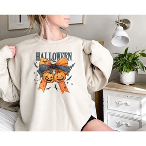 Puede incluir: Sudadera beige con un dise&ntilde;o de Halloween. El dise&ntilde;o presenta la palabra "HALLOWEEN" sobre un lazo con dos calabazas sonrientes, murci&eacute;lagos y estrellas. El lazo es negro y naranja.