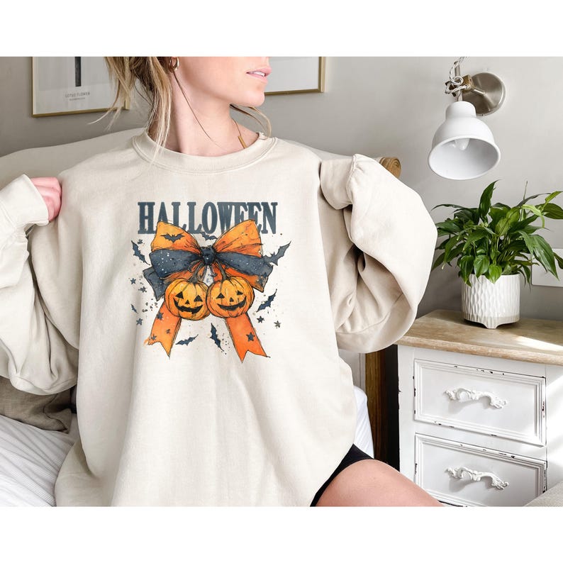 Puede incluir: Sudadera beige con un dise&ntilde;o de Halloween. El dise&ntilde;o presenta la palabra "HALLOWEEN" sobre un lazo con dos calabazas sonrientes, murci&eacute;lagos y estrellas. El lazo es negro y naranja.