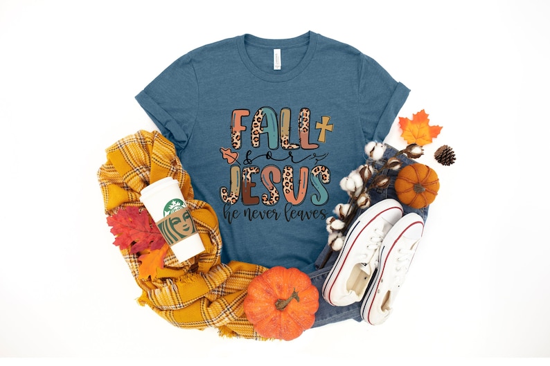 May include: A teal blue t-shirt with the text "Fall for Jesus he never leaves" in colorful letters with a leopard print design. The shirt is surrounded by fall-themed items like a plaid scarf, pumpkins, and fall leaves.