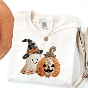 May include: Cream-coloured t-shirt with a Halloween design. The design features a smiling ghost in a witch hat and a jack-o'-lantern. A gold necklace with a pendant is draped across the shirt. The shirt is folded on a white surface.