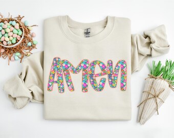 Puede incluir: Una sudadera beige con la palabra "AMEN" impresa en letras grandes y coloridas. Las letras están llenas de un patrón floral en tonos de rosa, azul, verde y amarillo.