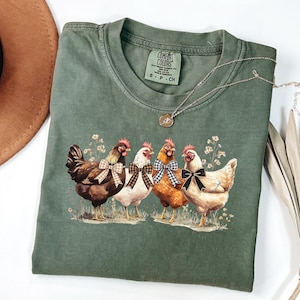 May include: Green Comfort Colors t-shirt featuring a graphic of four chickens with bow ties. The chickens are brown, white, and orange, standing on grass with small flowers. A silver necklace with a pendant is draped on the shirt.