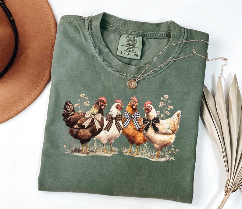 May include: Green Comfort Colors t-shirt featuring a graphic of four chickens with bow ties. The chickens are brown, white, and orange, standing on grass with small flowers. A silver necklace with a pendant is draped on the shirt.