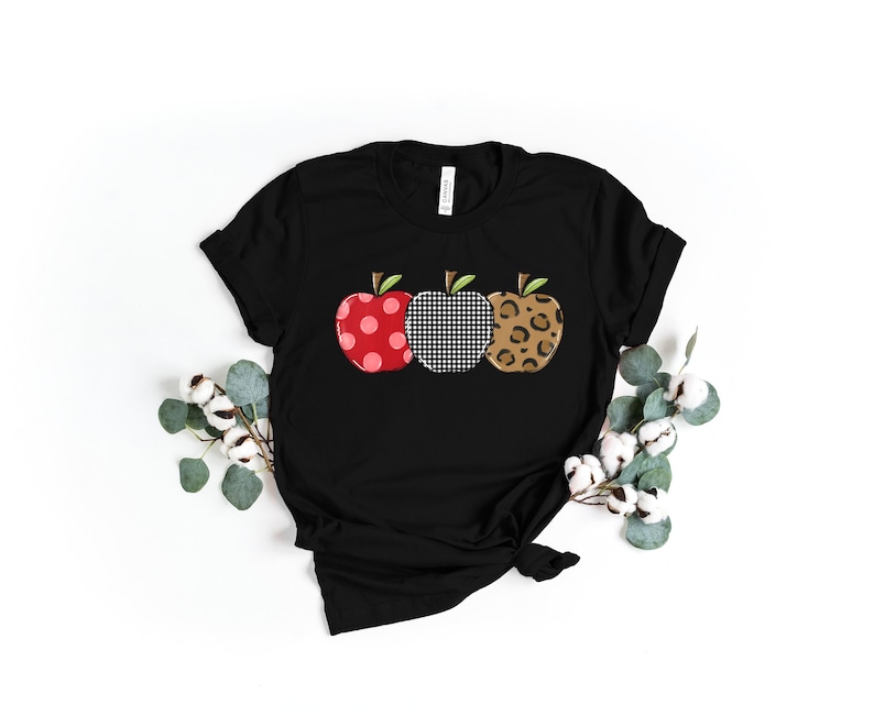 Pu&ograve; includere: T-shirt nero con tre disegni di mele. Le mele hanno motivi diversi: rosso a pois bianchi, quadretti bianchi e neri e stampa leopardata marrone. Le mele hanno foglie e steli verdi.