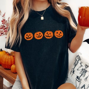 May include: A black t-shirt with a row of four orange jack-o'-lantern faces printed on the front. The jack-o'-lanterns have simple, smiling faces.