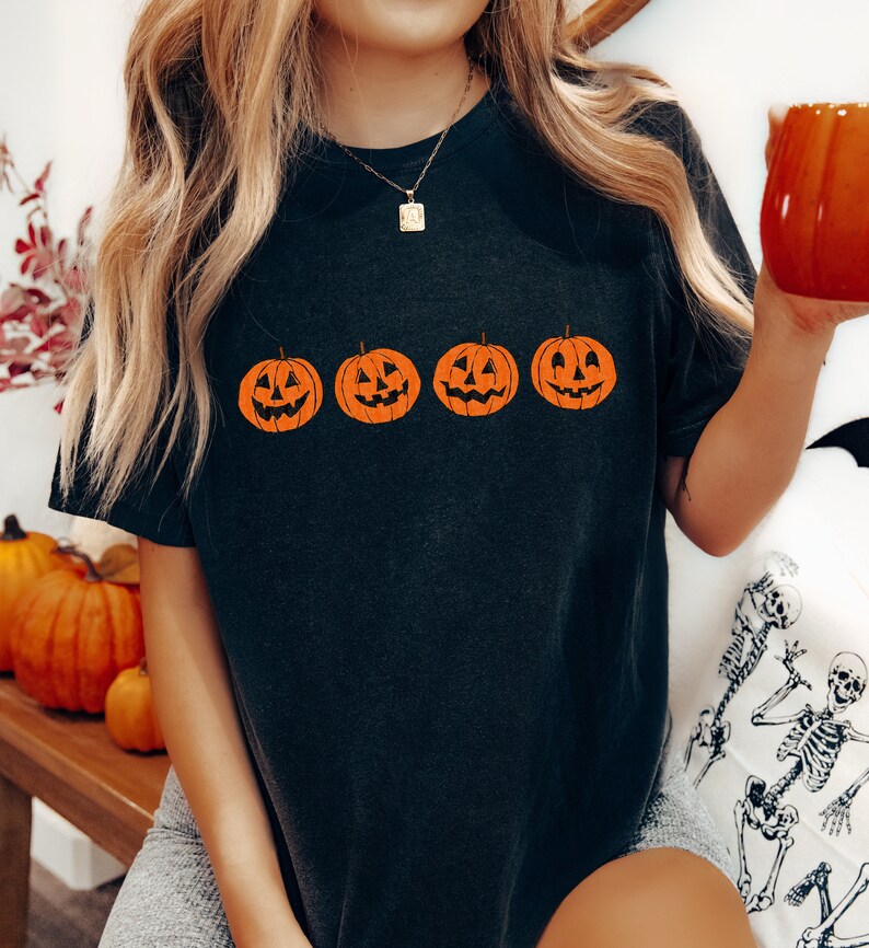 May include: A black t-shirt with a row of four orange jack-o'-lantern faces printed on the front. The jack-o'-lanterns have simple, smiling faces.