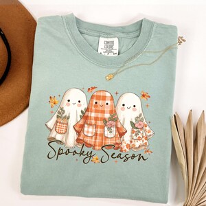 May include: A light green t-shirt featuring three cartoon ghosts with floral pockets and autumn leaves. The text "Spooky Season" is written in a script font below the ghosts. A gold necklace is draped across the shirt.