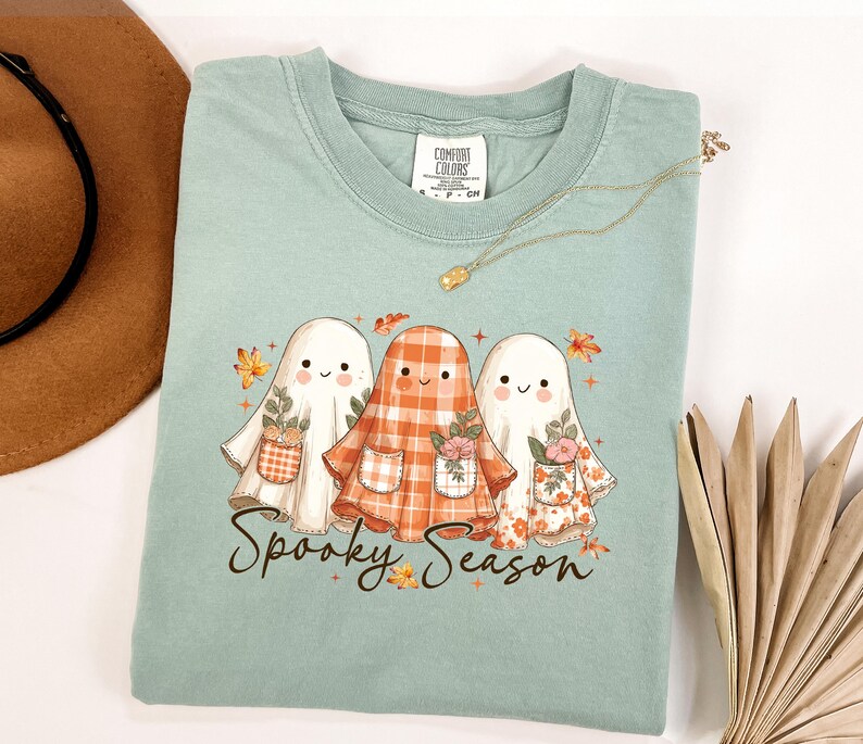 May include: A light green t-shirt featuring three cartoon ghosts with floral pockets and autumn leaves. The text "Spooky Season" is written in a script font below the ghosts. A gold necklace is draped across the shirt.