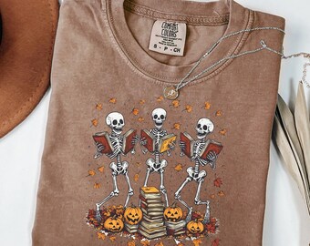 Puede incluir: Camiseta Comfort Colors marrón con un gráfico de tres esqueletos leyendo libros, rodeados de calabazas y hojas de otoño. Un collar plateado con un colgante está sobre la camiseta. Talla S.
