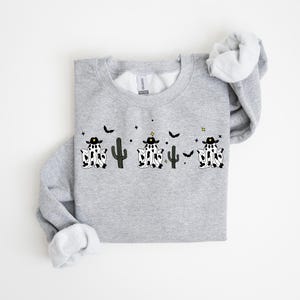 May include: A light gray sweatshirt featuring a whimsical design of three ghost figures dressed as cowboys, with black hats and cow-print bodies. The design includes black cacti, bats, and stars. The sweatshirt has a crew neck and long sleeves.