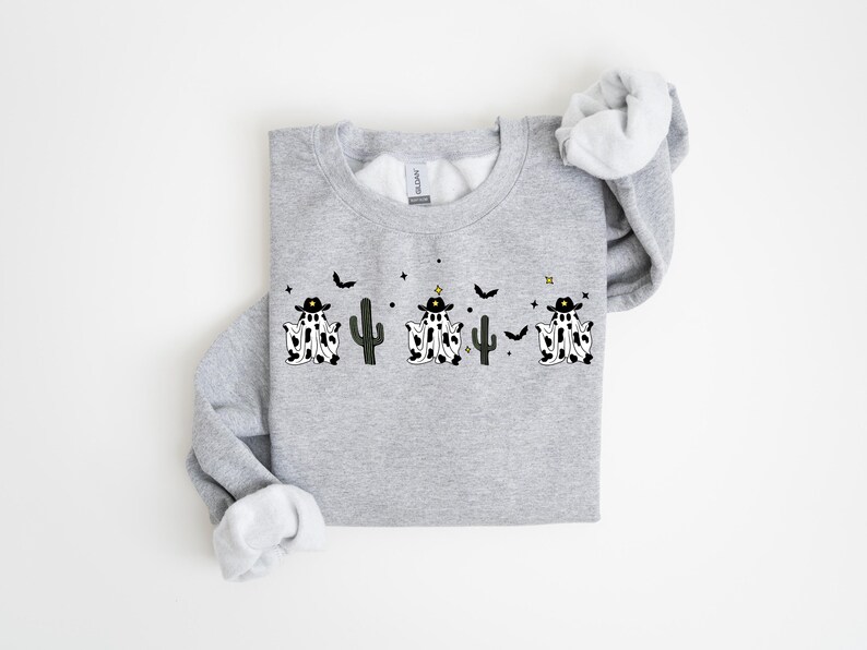 May include: A light gray sweatshirt featuring a whimsical design of three ghost figures dressed as cowboys, with black hats and cow-print bodies. The design includes black cacti, bats, and stars. The sweatshirt has a crew neck and long sleeves.