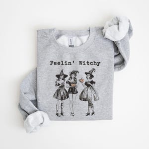 May include: Gray sweatshirt with the text "Feelin' Witchy" and a graphic of three witches in black dresses and pointy hats holding pumpkins.