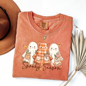 May include: A coral-colored t-shirt featuring a graphic of three cartoon ghosts with floral and plaid details. The text "Spooky Season" is below the ghosts. A gold necklace and a brown hat are also visible.