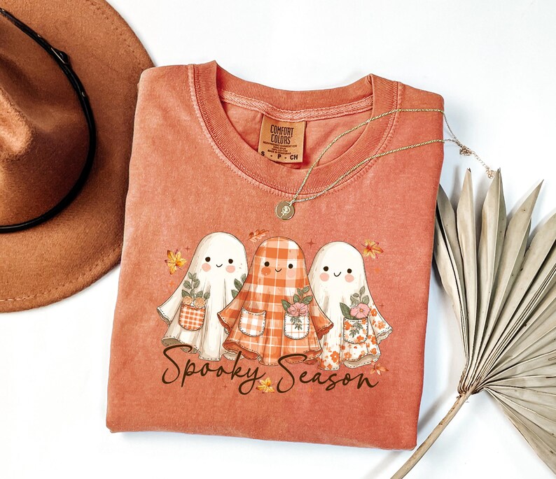 May include: A coral-colored t-shirt featuring a graphic of three cartoon ghosts with floral and plaid details. The text "Spooky Season" is below the ghosts. A gold necklace and a brown hat are also visible.