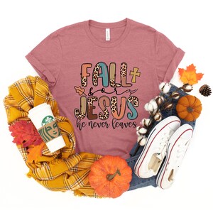 May include: A pink t-shirt with the text "Fall + Jesus he never leaves" in colorful letters with a leopard print design. The shirt is surrounded by fall-themed items like pumpkins, leaves, and a plaid scarf.