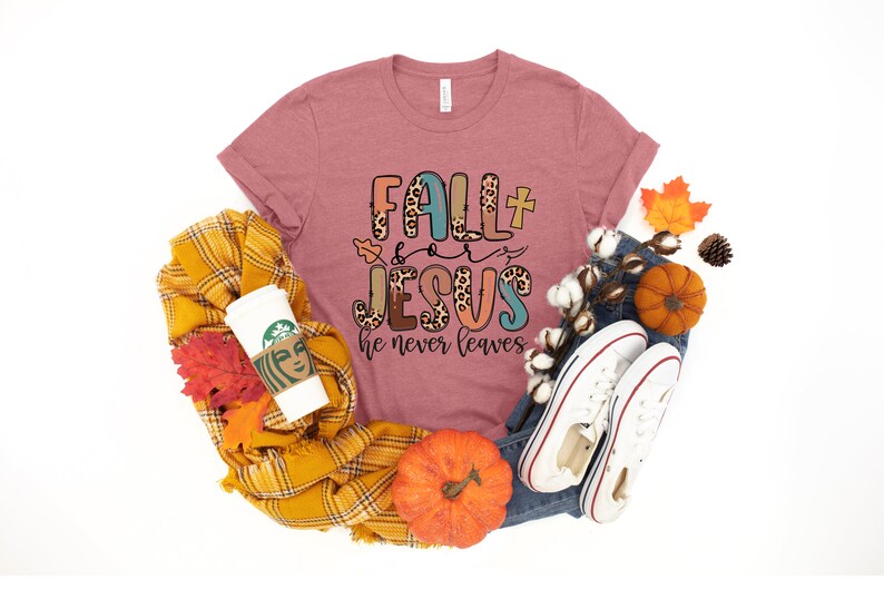 May include: A pink t-shirt with the text "Fall + Jesus he never leaves" in colorful letters with a leopard print design. The shirt is surrounded by fall-themed items like pumpkins, leaves, and a plaid scarf.