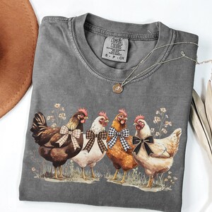 May include: A gray Comfort Colors t-shirt featuring a graphic of four chickens with bowties. The chickens are brown, white, and orange, standing in a field of grass and flowers. A silver necklace and a brown hat are also visible.