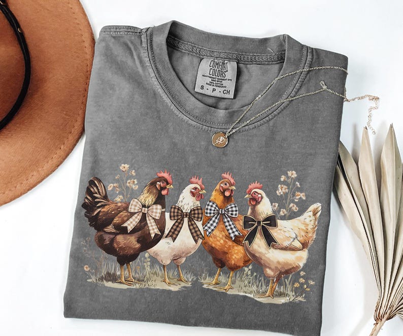May include: A gray Comfort Colors t-shirt featuring a graphic of four chickens with bowties. The chickens are brown, white, and orange, standing in a field of grass and flowers. A silver necklace and a brown hat are also visible.