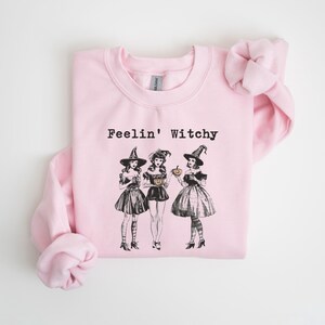 May include: Pink sweatshirt with the text "Feelin' Witchy" and a graphic of three witches in black dresses and pointy hats holding pumpkins.