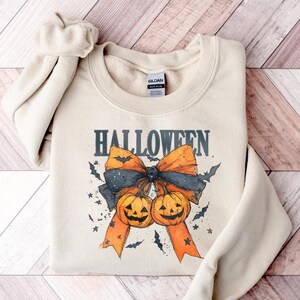 Puede incluir: Sudadera beige con un dise&ntilde;o de Halloween. El dise&ntilde;o presenta la palabra "HALLOWEEN" sobre un gran lazo con cintas naranjas y un centro azul oscuro. Tambi&eacute;n se incluyen dos calabazas y murci&eacute;lagos.